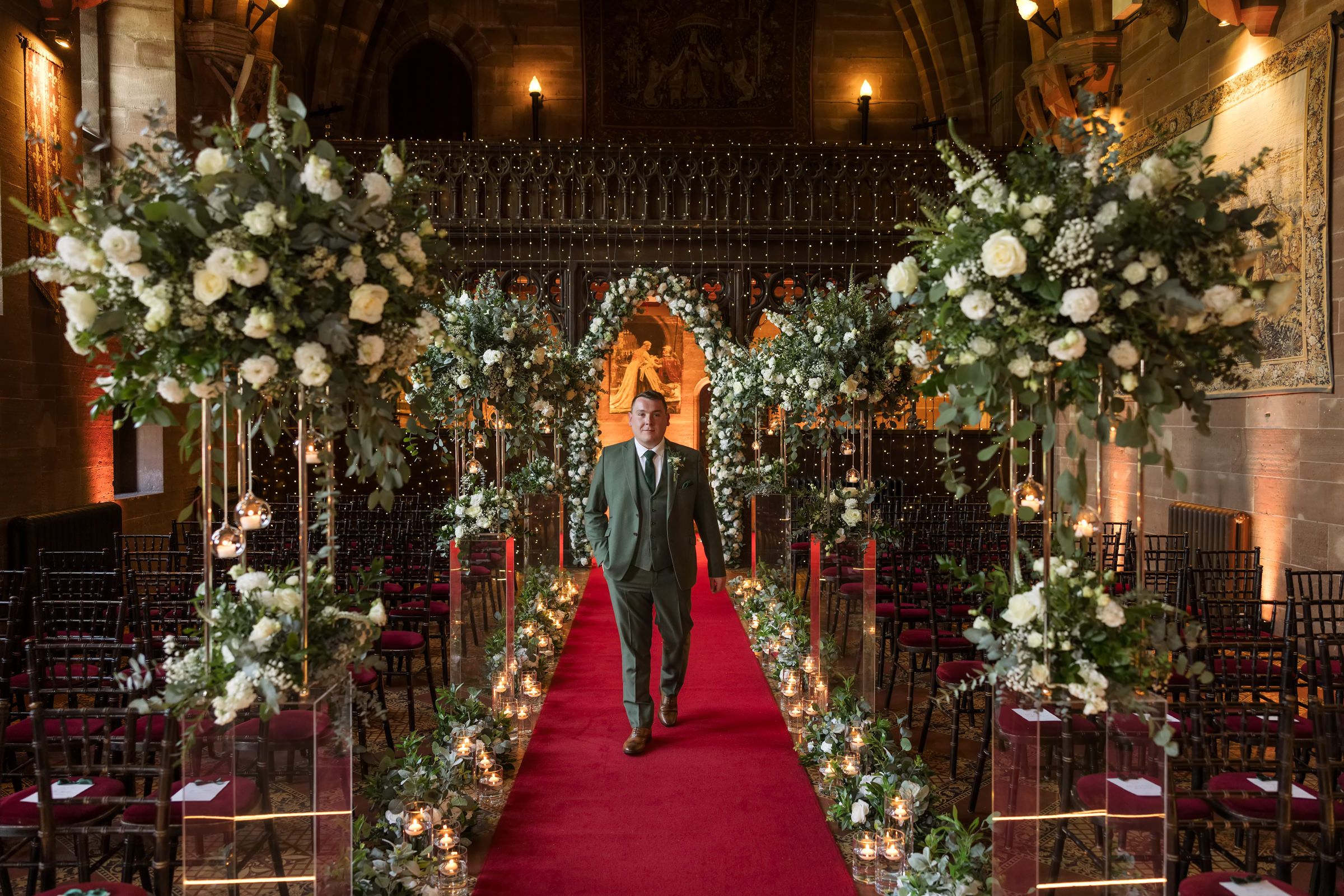 Elegant wedding aisle with floral arches of white roses and greenery lining a red carpet in a grand hall.