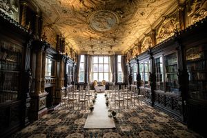 Ornate library wedding venue with gilded ceiling, dark wood bookshelves, and chairs arranged along a floral aisle runner.