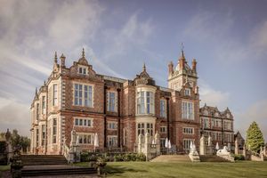 Grand Victorian red-brick manor house with ornate architecture, turrets, and manicured lawns under a blue sky.