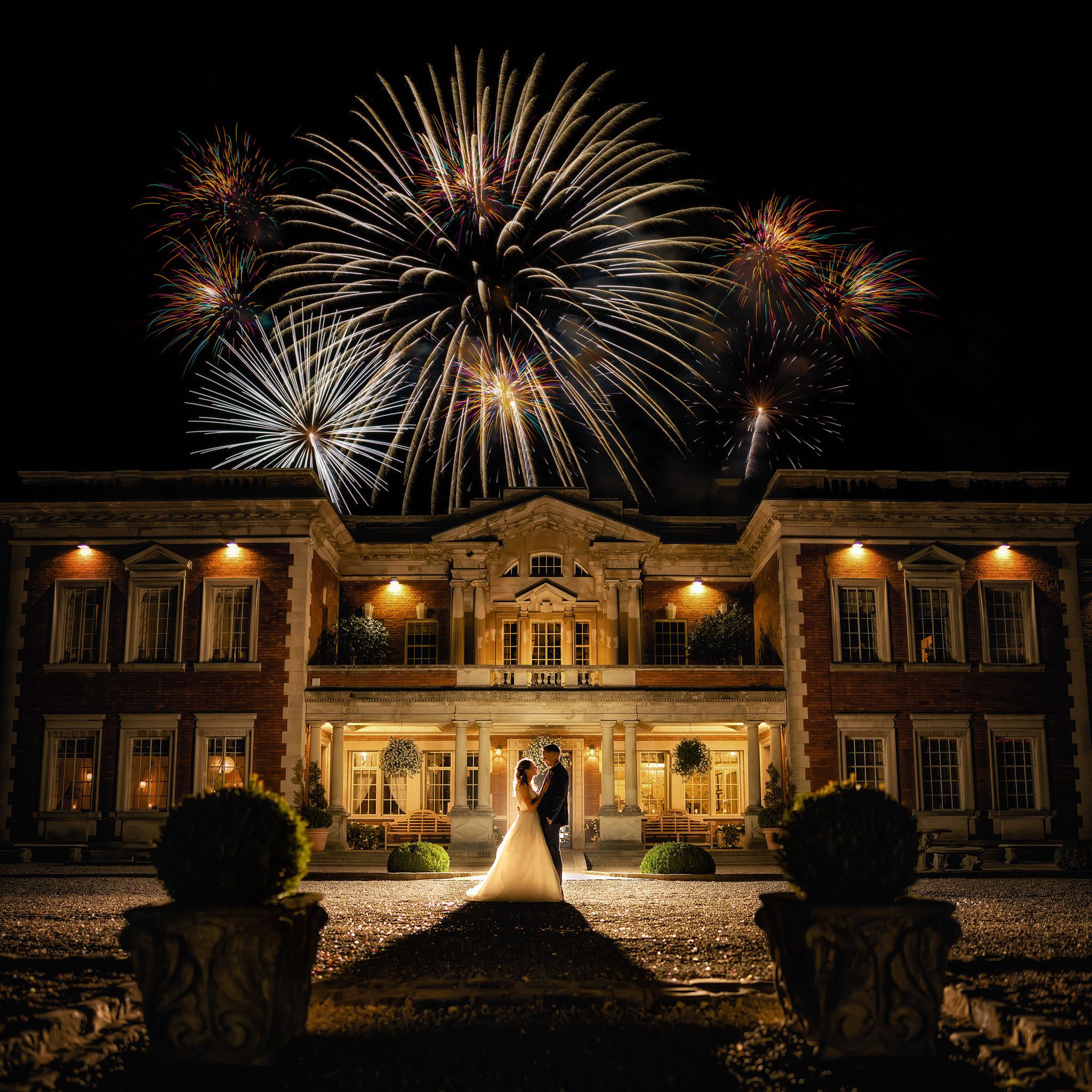 Bride and groom silhouetted before a grand illuminated manor house with stunning fireworks display lighting the night sky.