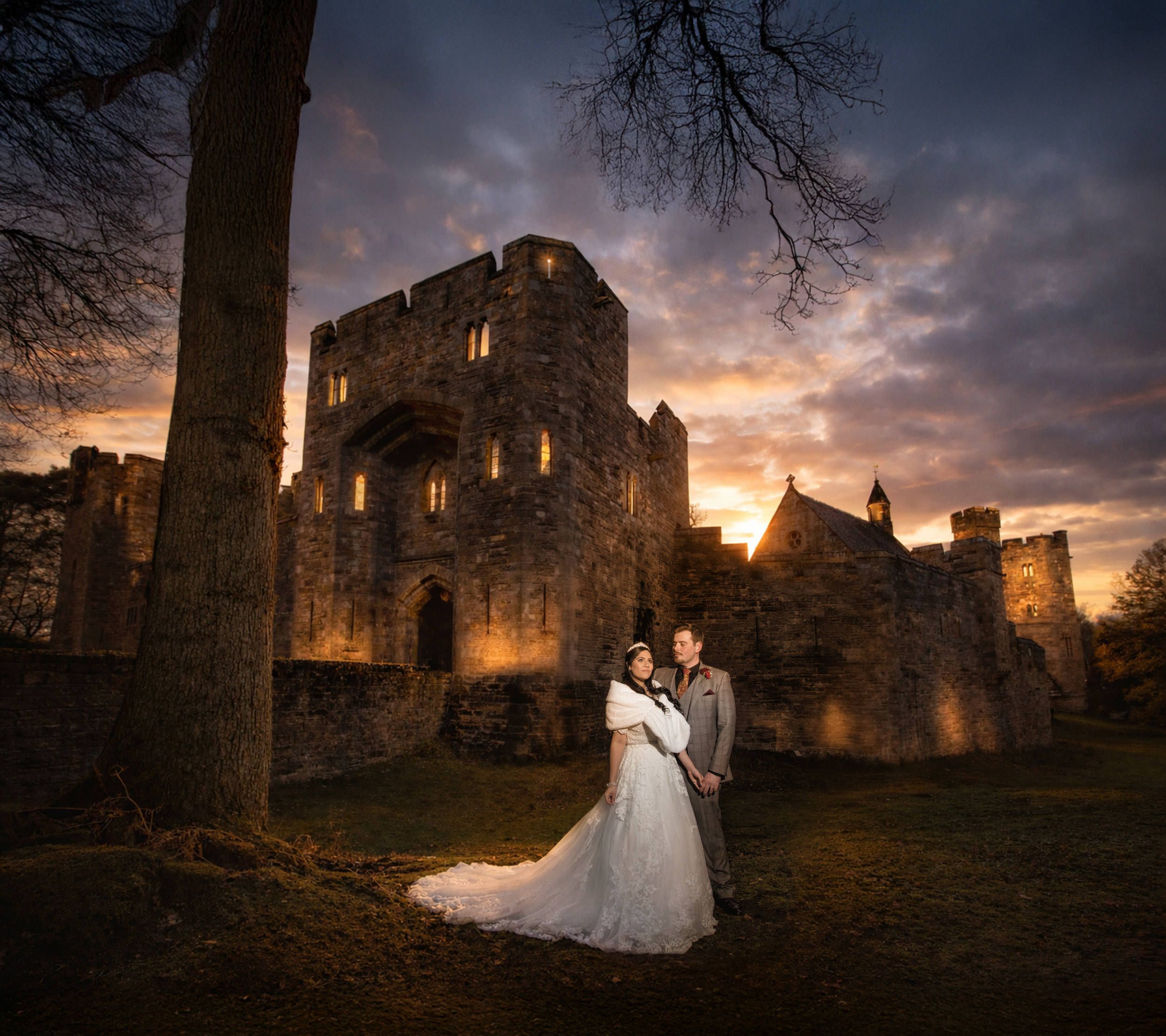 Bride and groom embrace at sunset before an illuminated stone castle with dramatic cloudy sky.