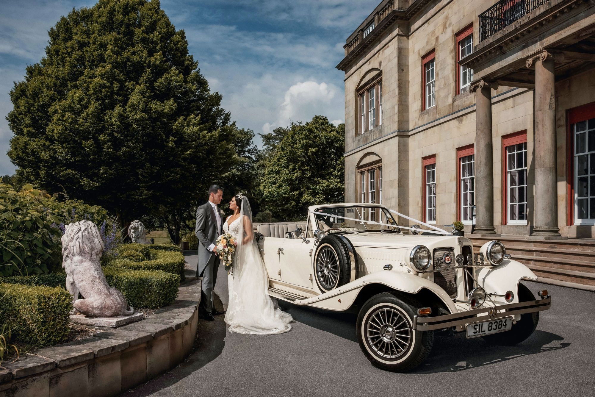 Bride and groom pose beside a vintage white wedding car outside an elegant manor house venue.
