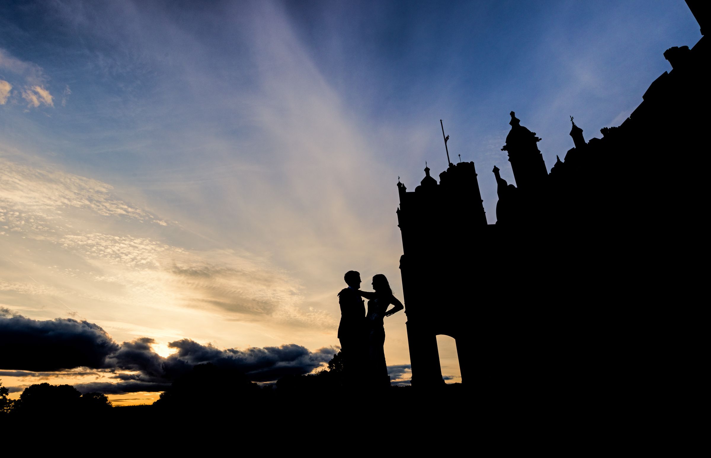 Silhouette of a couple embracing atop a castle battlement at sunset with dramatic rays of light.