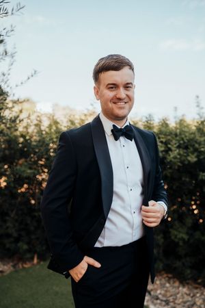 A person in a black tuxedo and white bowtie poses outdoors at sunset with a joyful expression.