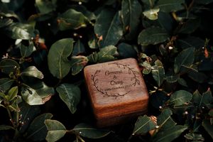 Small wooden ring box nestled among dark green leaves and foliage in natural outdoor lighting.