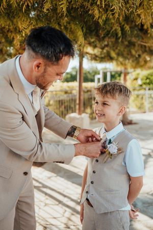A tender moment at a wedding as a man in a beige suit helps a young boy with a light blue boutonniere.