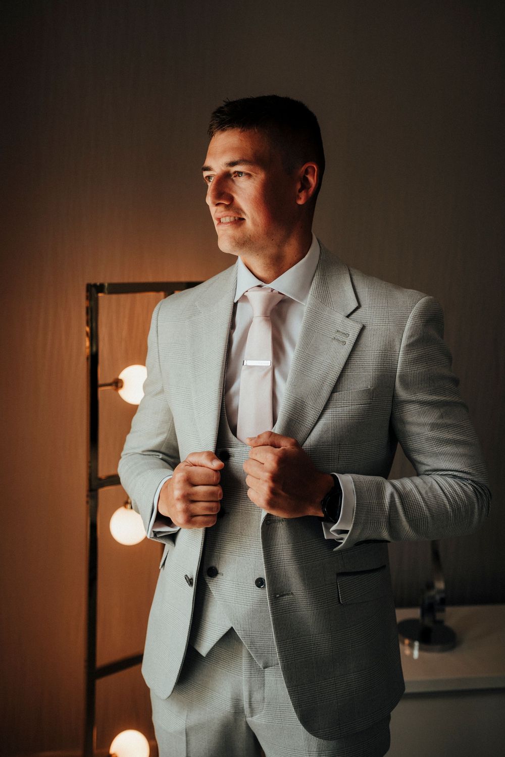 A man in a light gray suit adjusts his tie while getting ready in a dimly lit room with vanity lights.