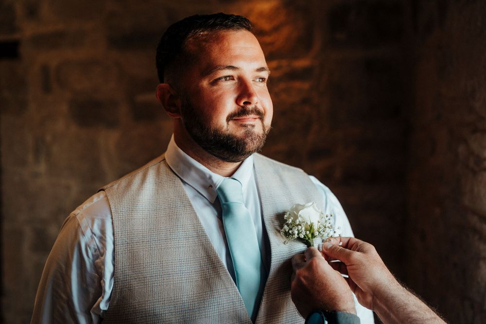 A person in a light gray suit and mint tie adjusts their boutonniere against a rustic stone wall background.