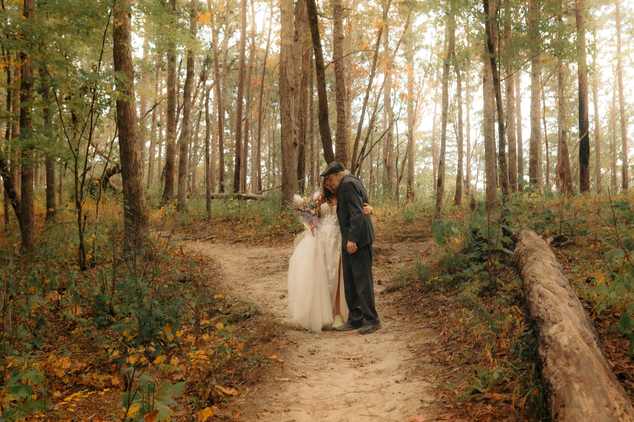 Bride and groom share a romantic kiss on a forest path surrounded by tall trees and golden autumn foliage.
