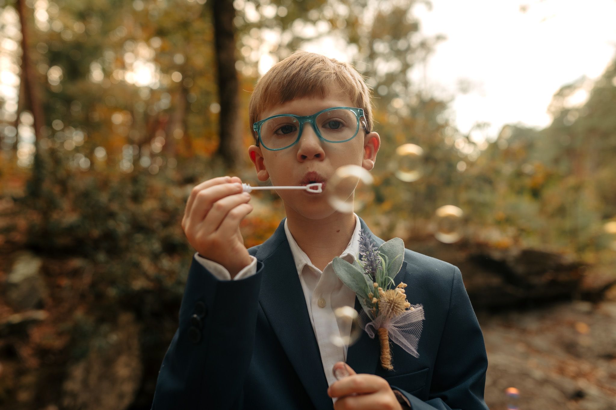 Child in formal attire blowing bubbles in outdoor setting.