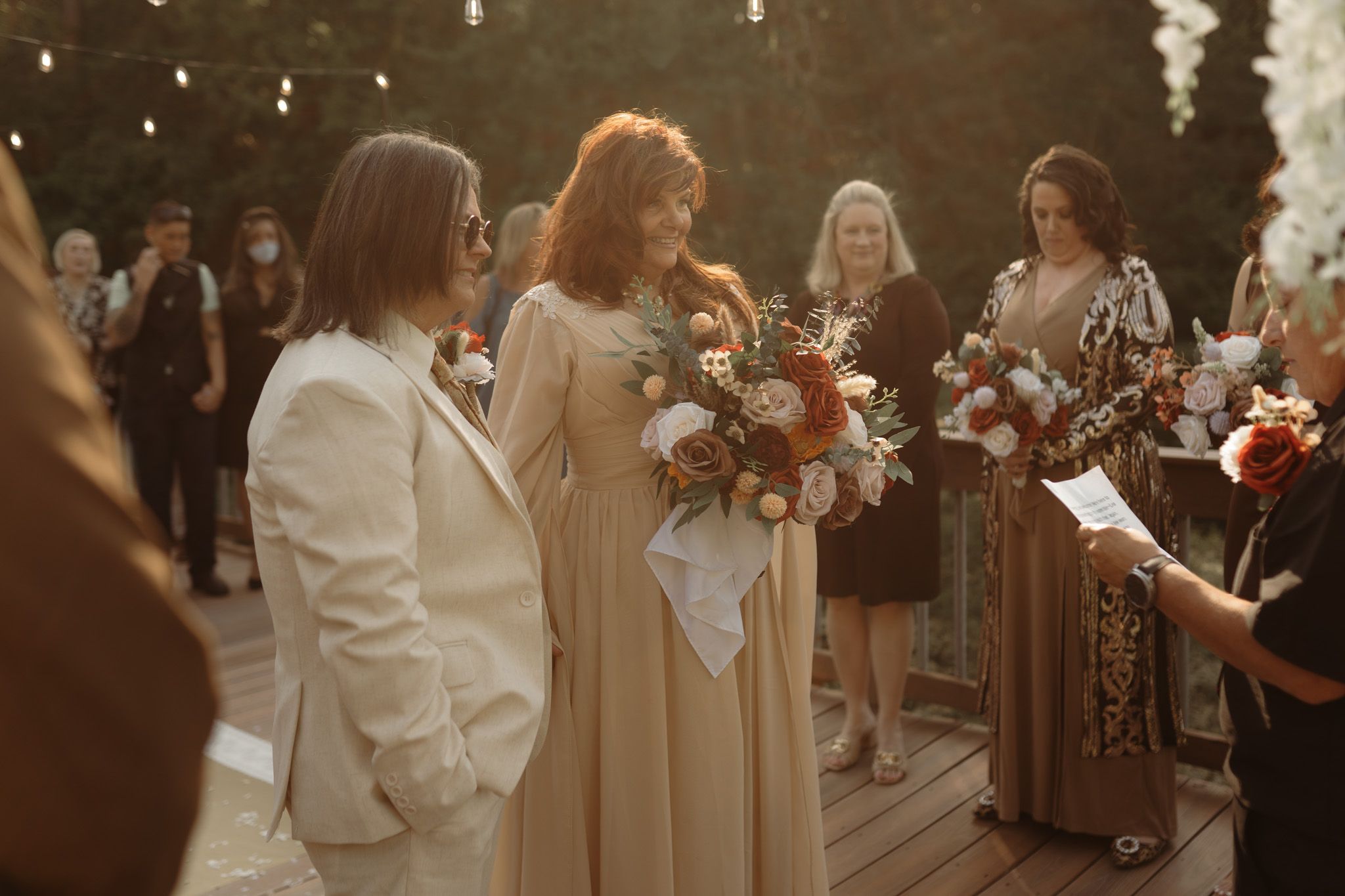 Two brides stand together at outdoor wedding ceremony, one in white suit, other in beige gown holding floral bouquet.