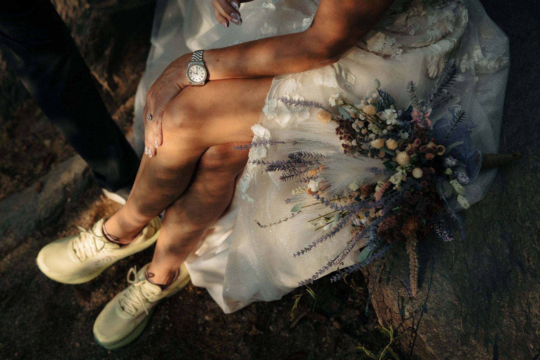 Bride sitting on rocks in white dress with wildflower bouquet and sneakers beside groom wearing a watch.