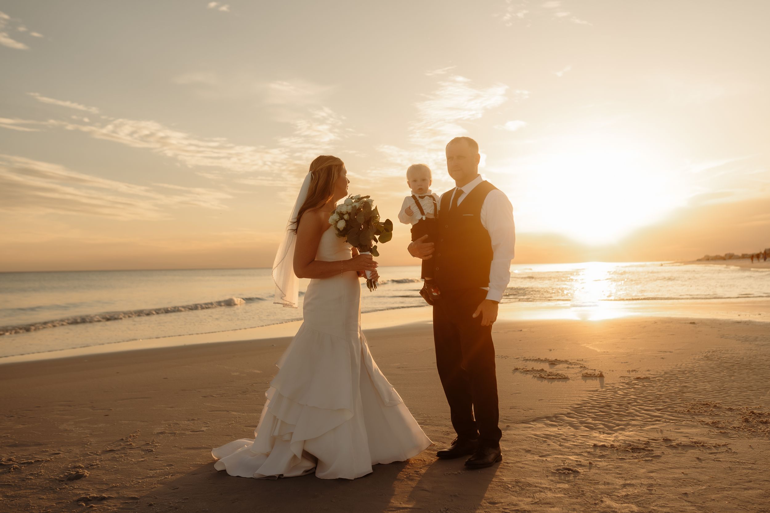 Bride and groom stand on a beach at golden sunset, bride holding bouquet, with a child between them.