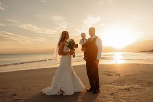 Bride and groom stand on a beach at golden sunset, bride holding bouquet, with a child between them.