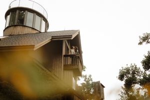 Couple standing on a wooden balcony of a rustic building with a glass tower, surrounded by trees.