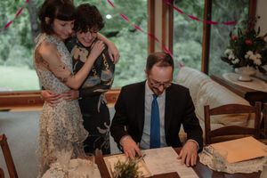 Man in suit signing documents at table while woman holds child during an intimate wedding ceremony indoors.