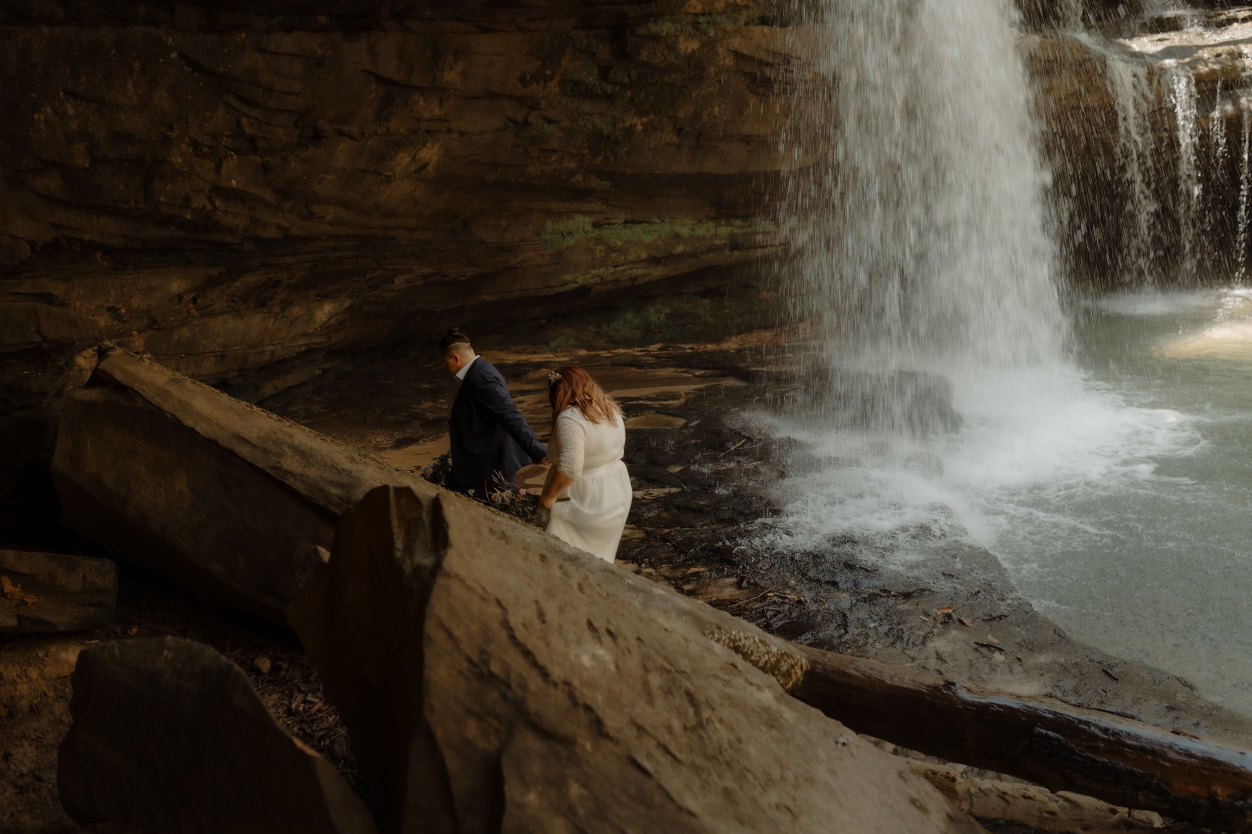A couple sits together on a rocky ledge overlooking a dramatic waterfall in a cave-like setting.