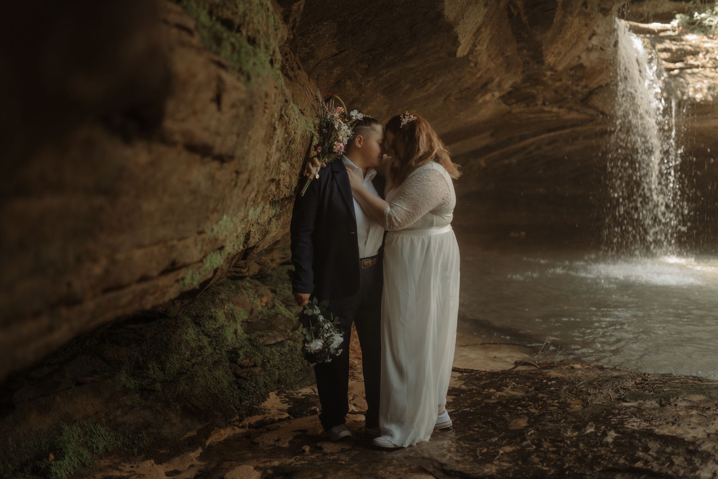 A romantic moment captured under a rocky overhang with a waterfall in the background.