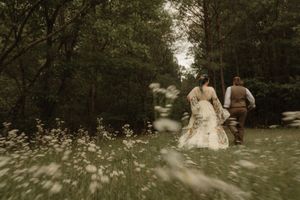A bride and groom walk hand in hand through a wildflower meadow surrounded by lush green trees.