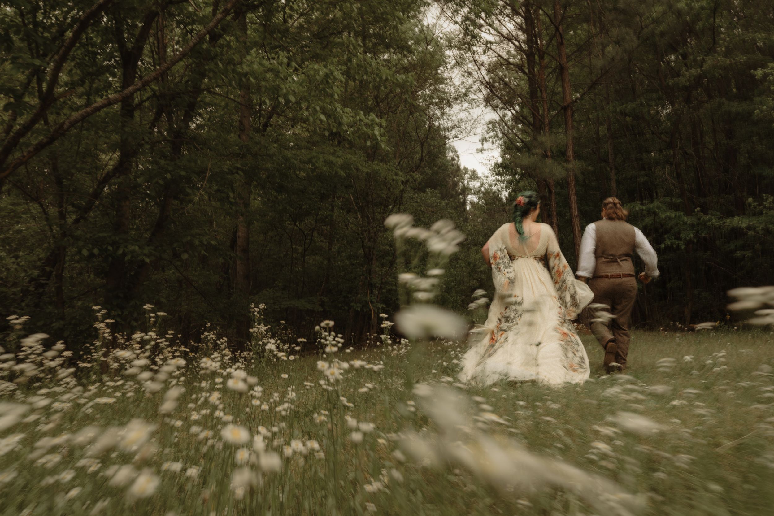A bride and groom walk hand in hand through a wildflower meadow surrounded by lush green trees.