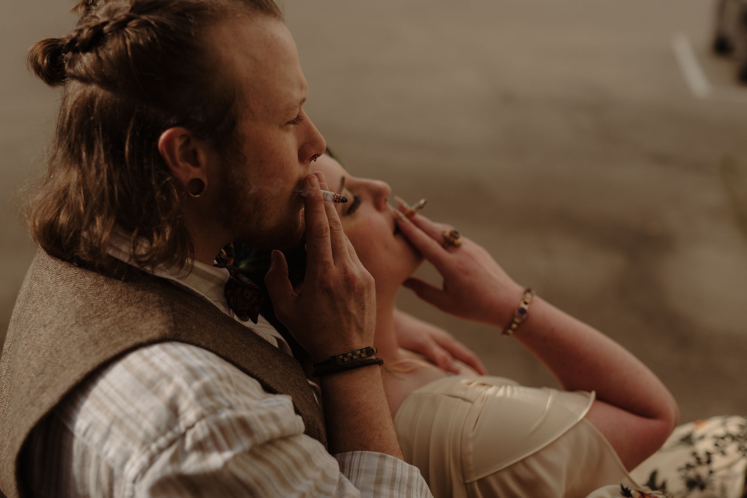 Man with long hair and vest smoking a cigarette while relaxing indoors with a woman wearing a bracelet.