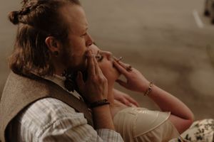 Man with long hair and vest smoking a cigarette while relaxing indoors with a woman wearing a bracelet.