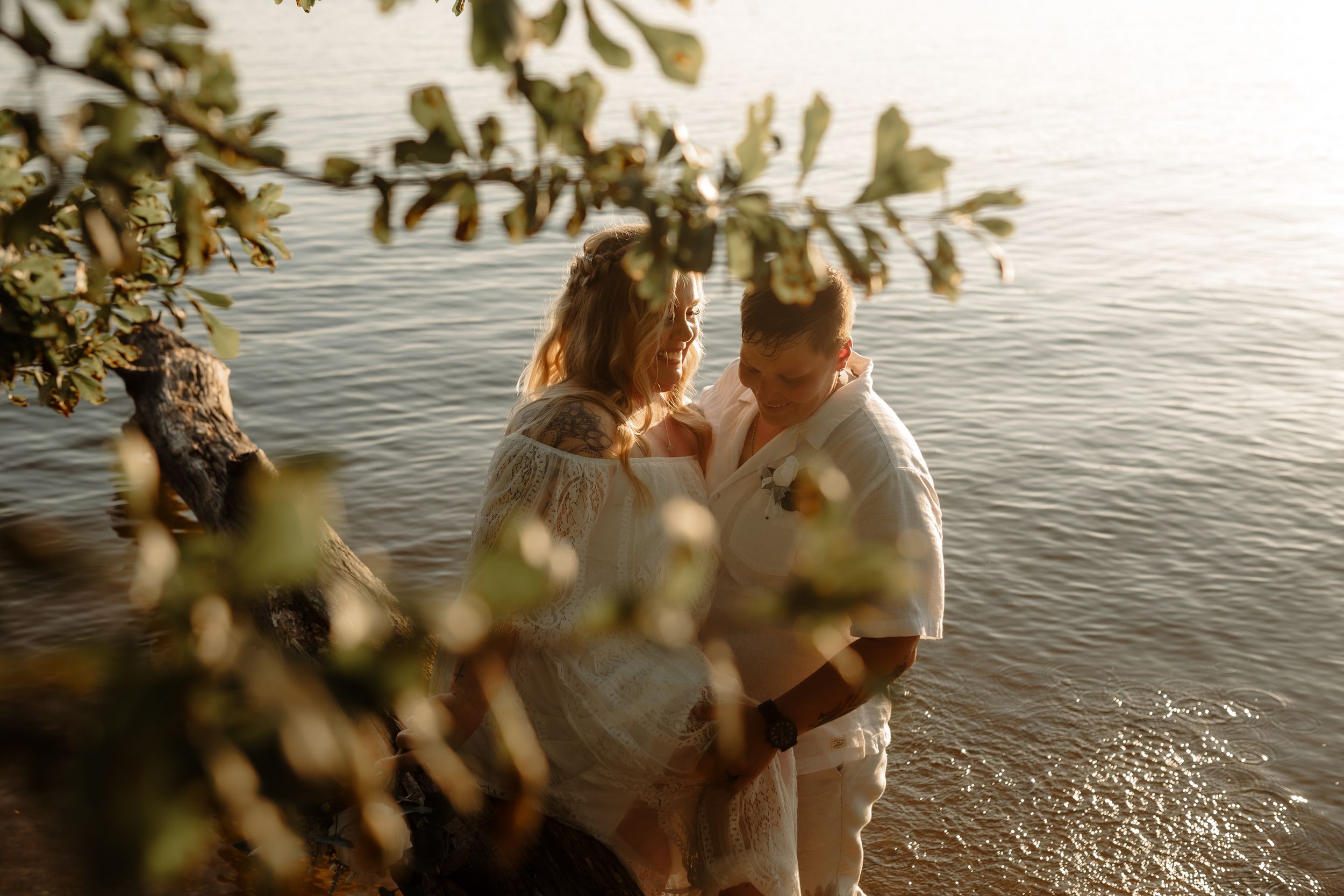 A romantic couple embraces by the waterfront at sunset, framed by leafy branches in the foreground.