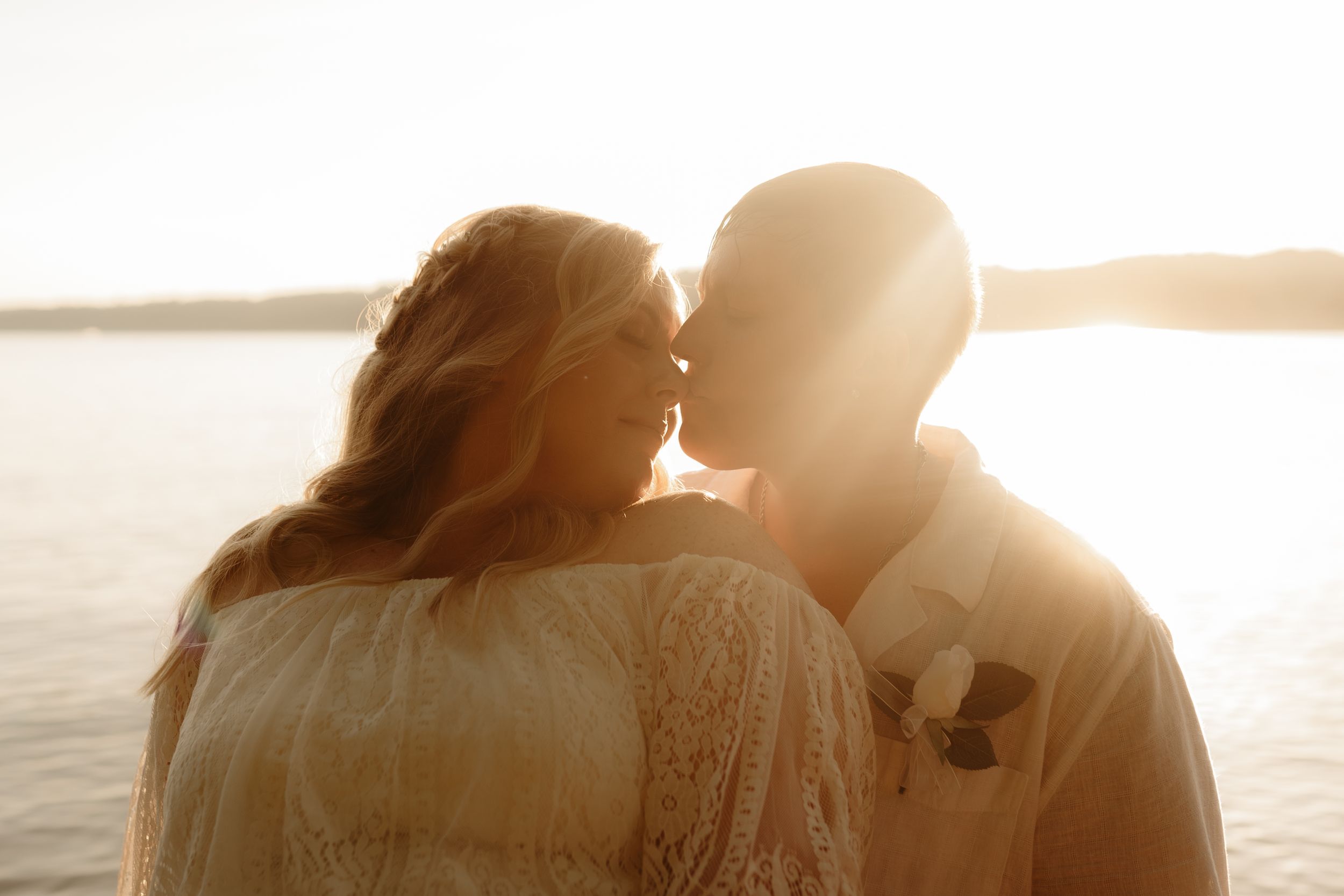 A romantic couple shares an intimate moment at sunset by the water in soft golden light.
