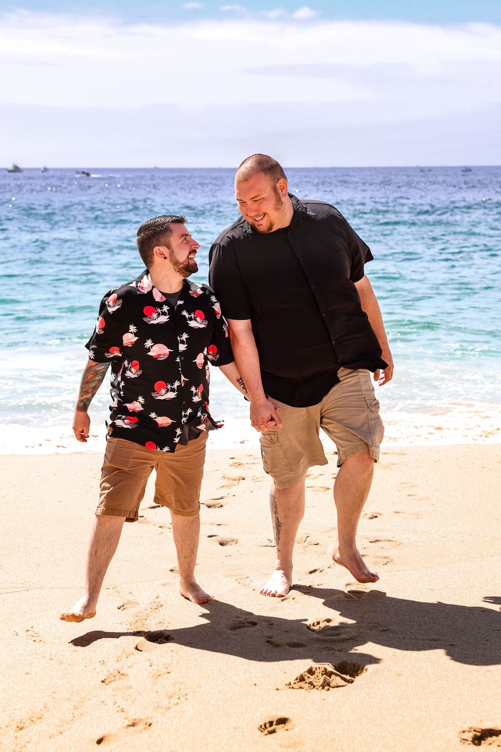 Two people walking and laughing together on a sunny beach with blue ocean in background.