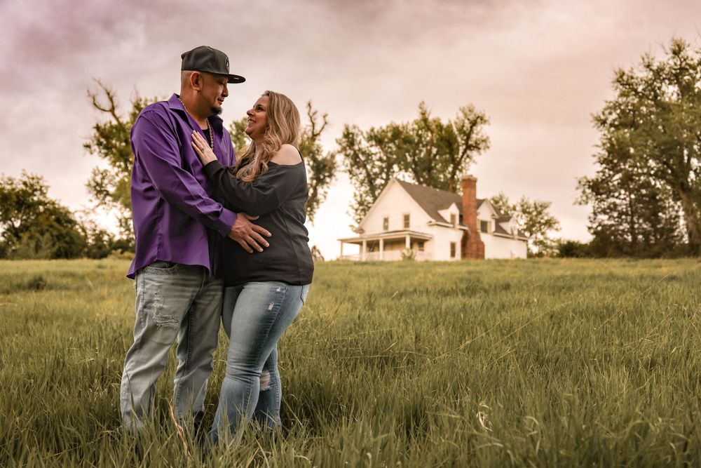 A couple shares an intimate moment in a field with a farmhouse in the background at sunset.