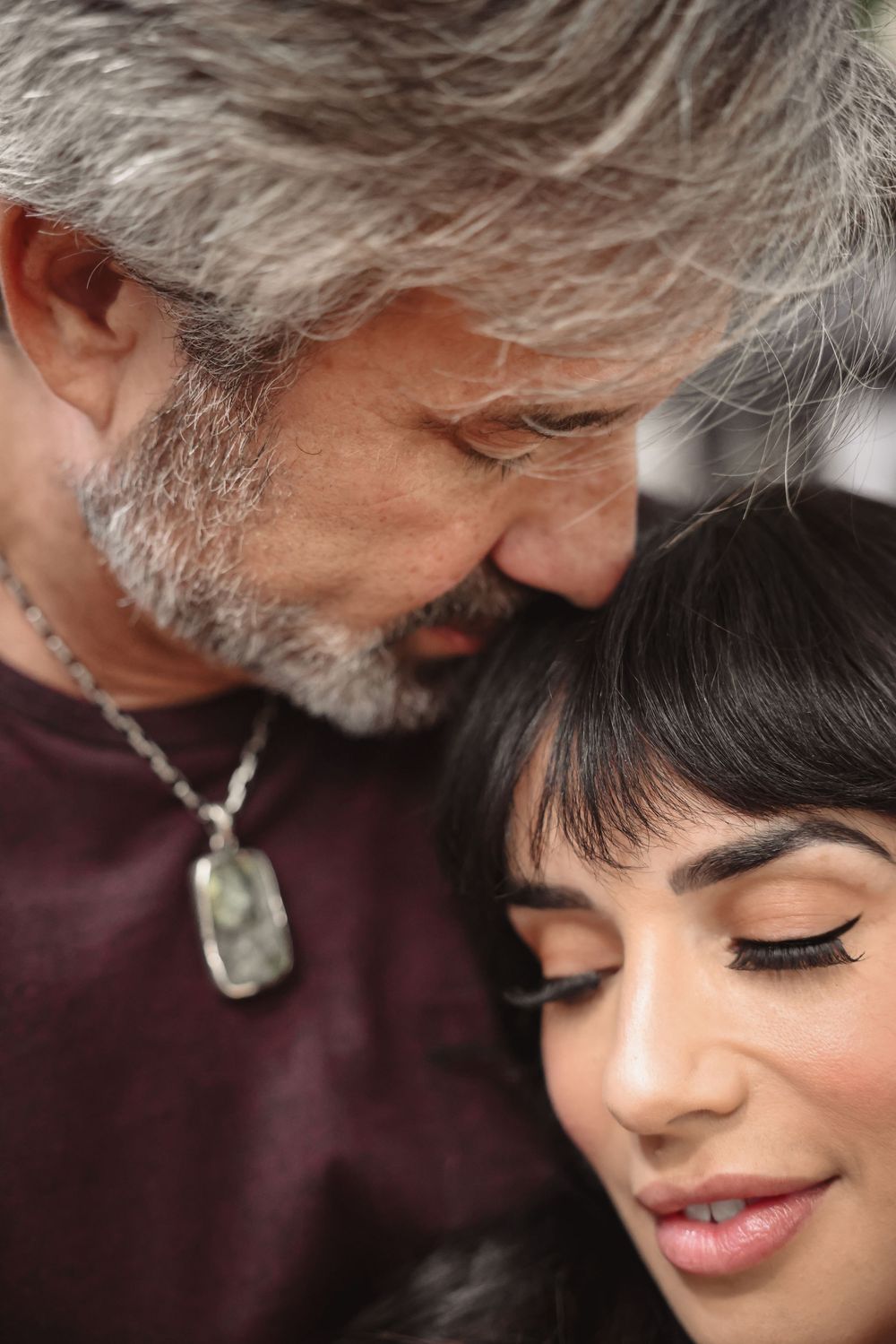 An intimate close-up portrait of two people in a tender moment, with one wearing a burgundy top and silver pendant.