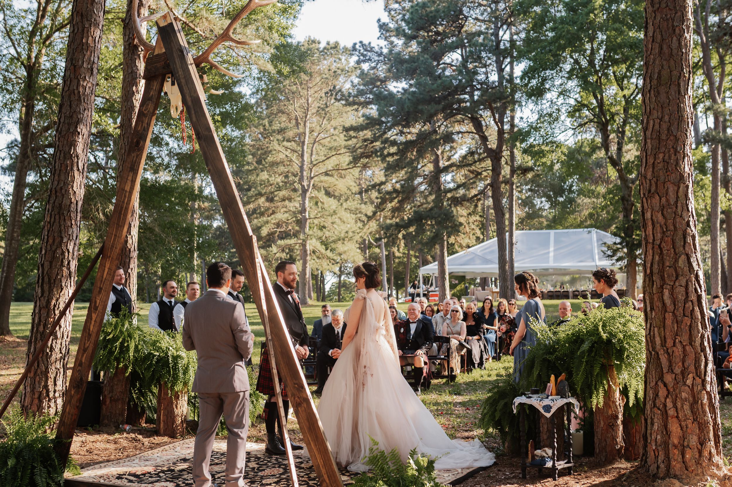 Outdoor woodland wedding ceremony with couple at a wooden arch altar surrounded by pine trees and seated guests.