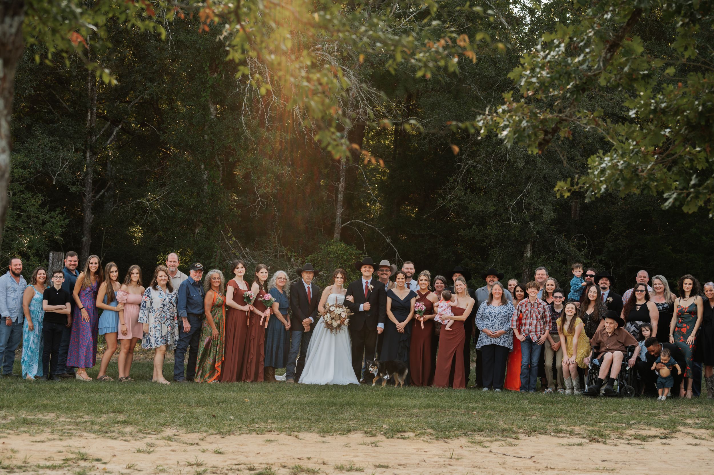 Large wedding party group photo on a grassy field surrounded by autumn trees in golden evening light.