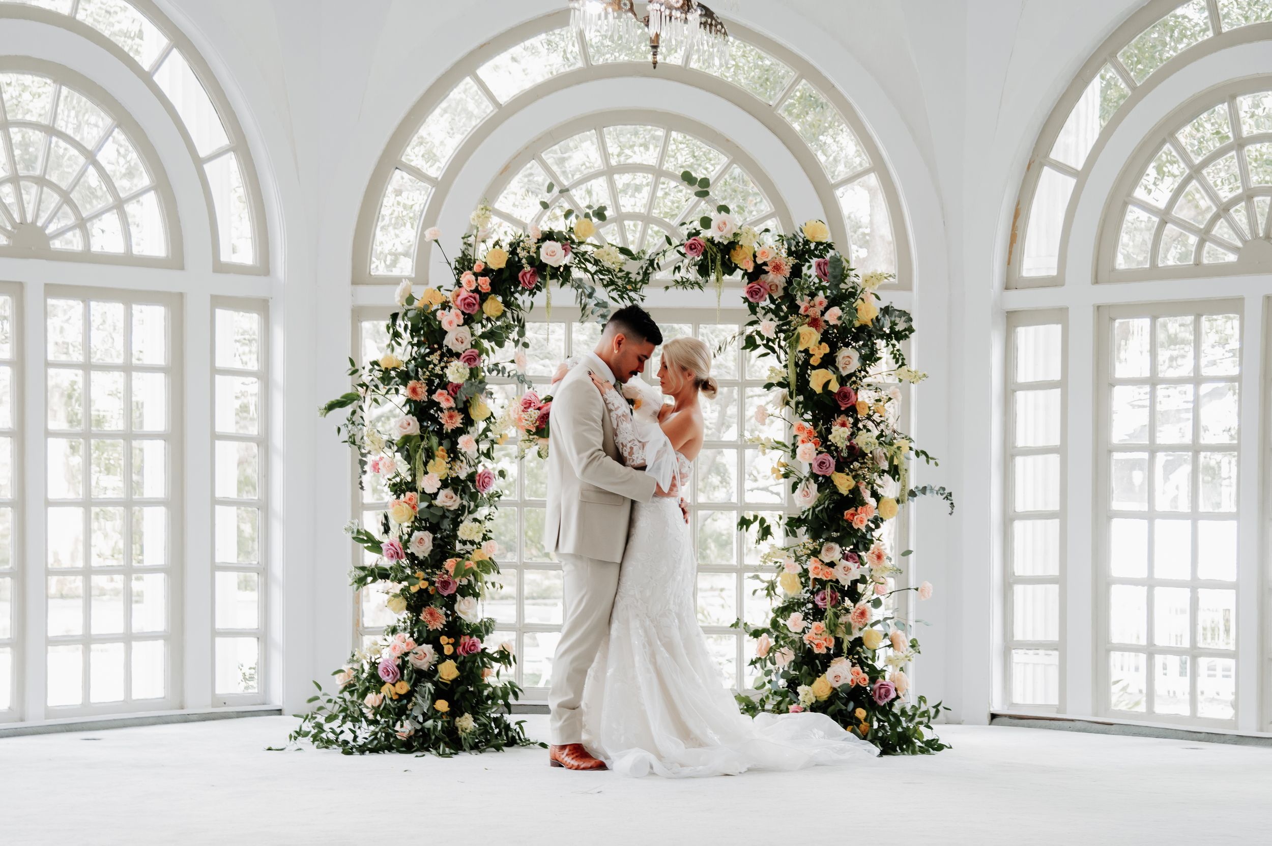 Couple shares a romantic embrace under a colorful floral arch in a bright white sunlit ballroom.