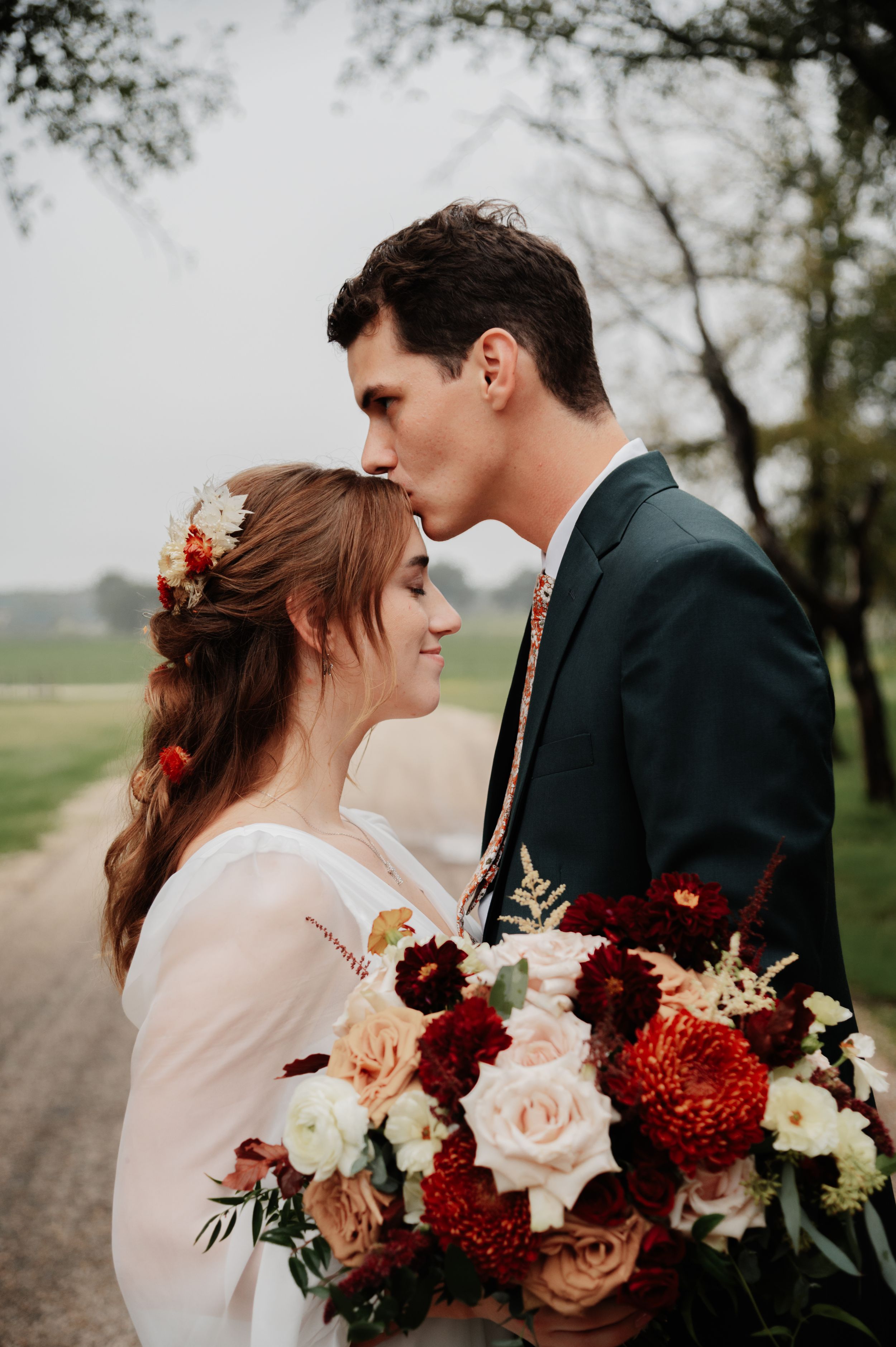 Groom tenderly kisses bride on forehead as she smiles holding a lush red and blush floral bouquet.