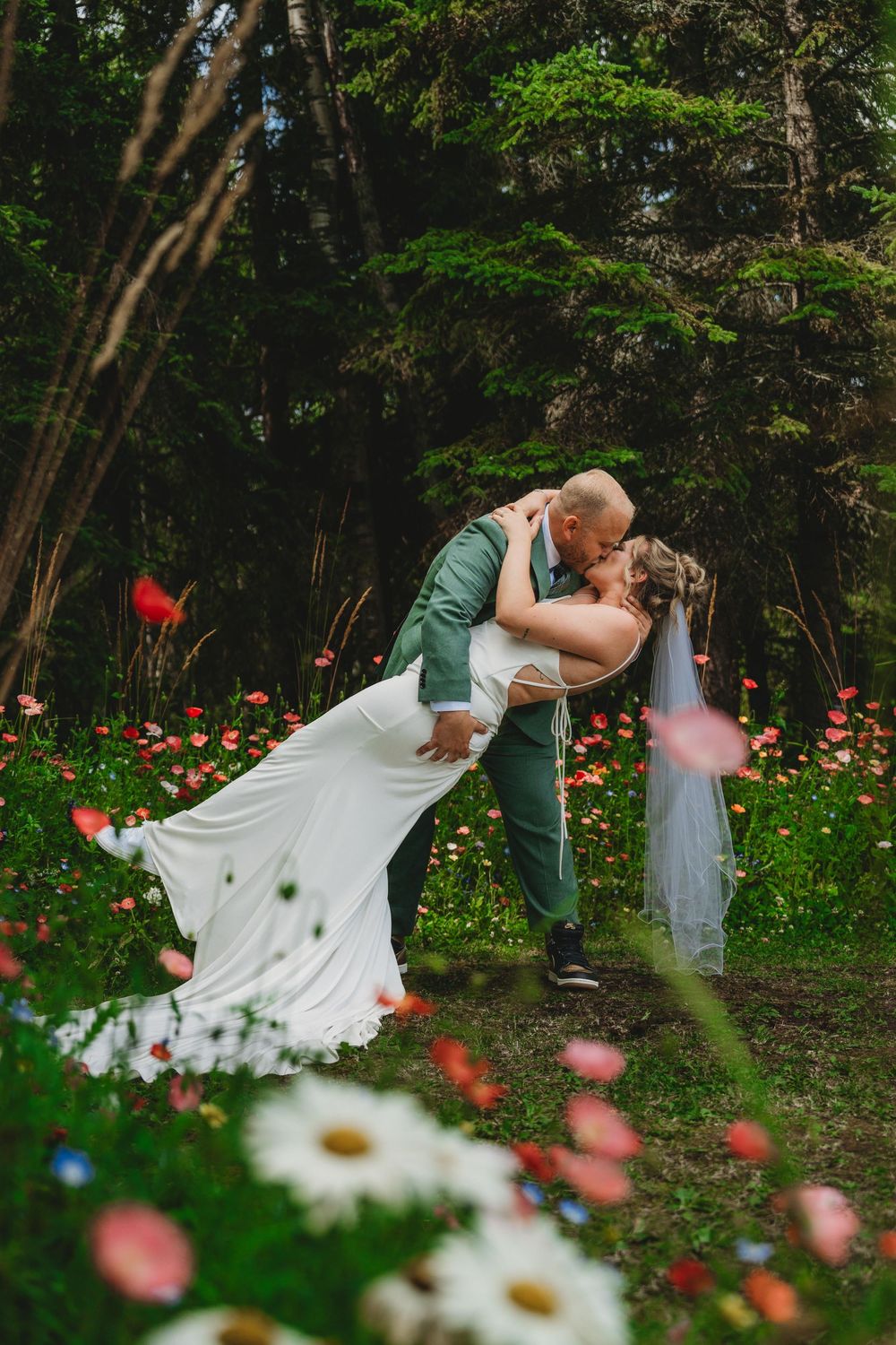 Bride and groom share a romantic dip kiss in a wildflower meadow surrounded by lush green forest.