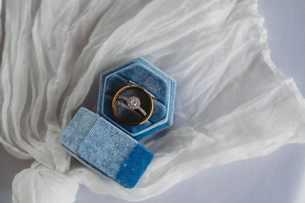 Gold diamond engagement ring displayed in a blue hexagonal velvet ring box on white fabric.