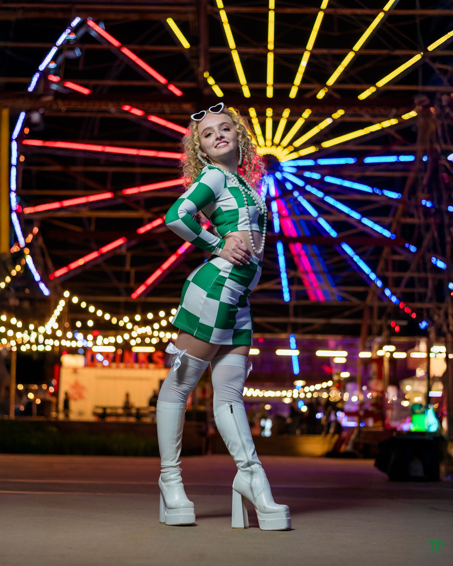 Person in green checkered dress and white boots poses in front of colorful illuminated carnival ferris wheel at night.