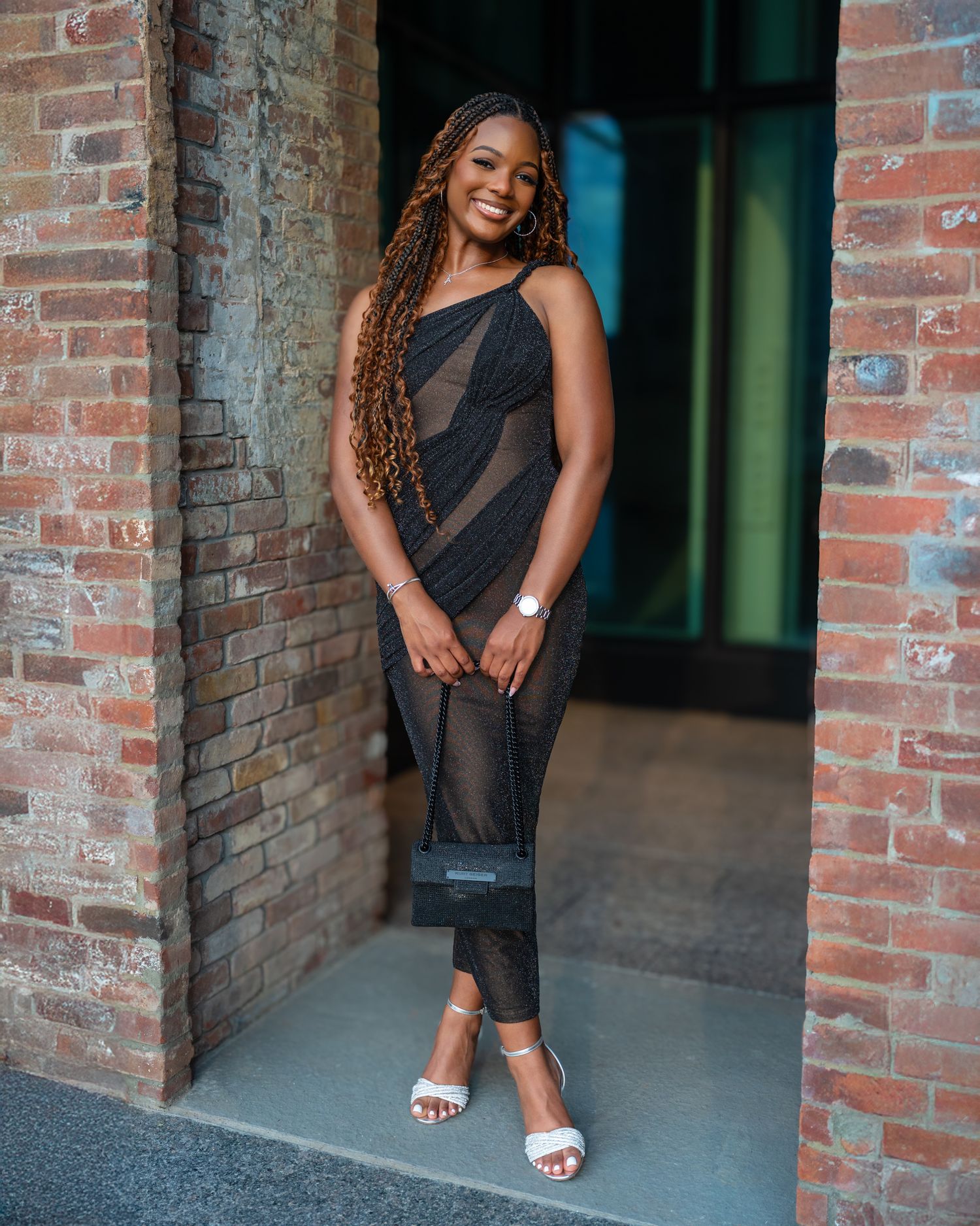 A confident woman in a black dress poses against exposed brick walls in an urban photoshoot setting.