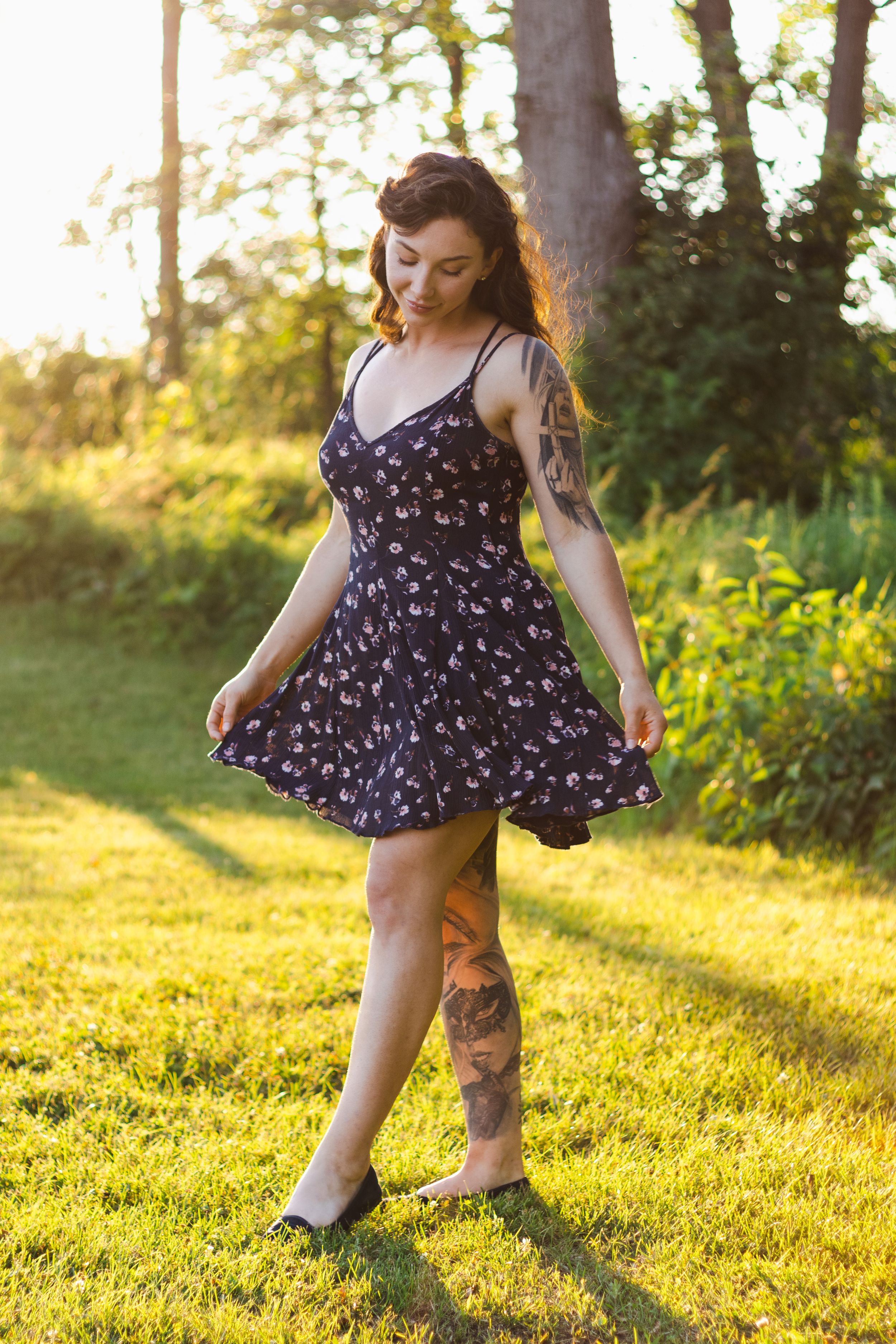 A floral summer dress twirls in golden sunlight as tattooed legs dance on lush green grass at sunset.