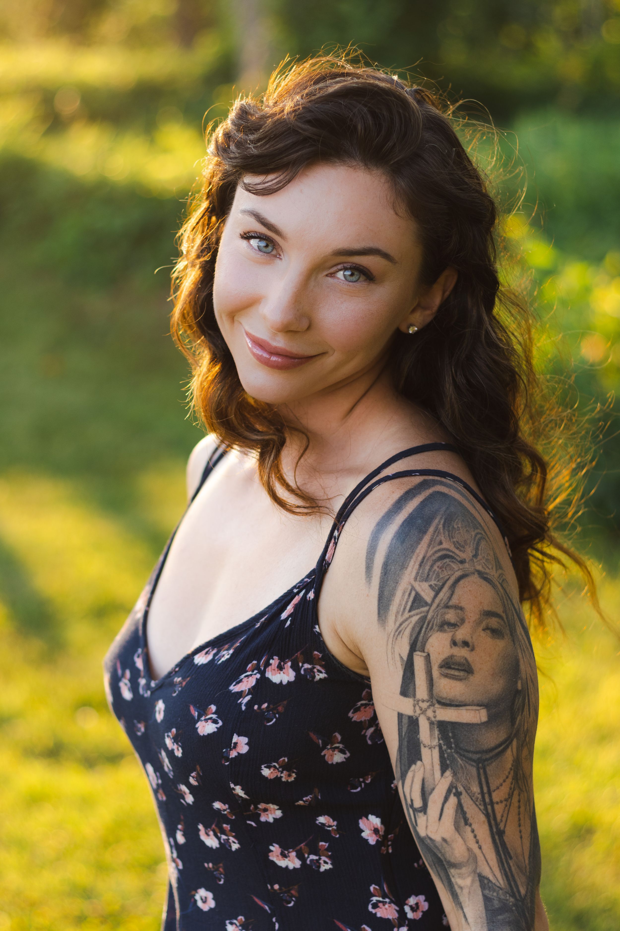 Portrait of a woman in a floral dress with dark wavy hair and arm tattoo standing in golden sunlit garden.