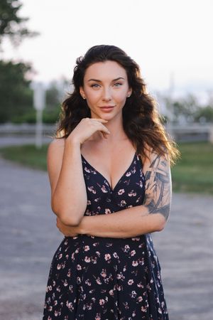 Woman wearing a navy floral dress poses outdoors with hand on chin and detailed arm tattoo.