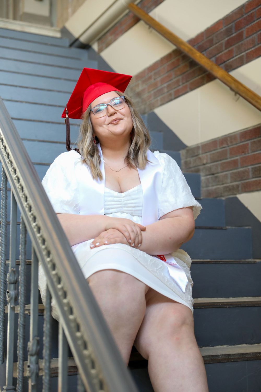 Graduate in red cap and gown sitting on outdoor stairs with brick wall background.
