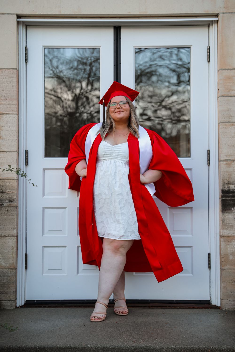 Graduate in red cap and gown poses confidently in front of white double doors.