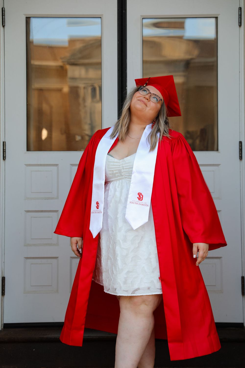 Graduate in red regalia leans back joyfully against white doors.