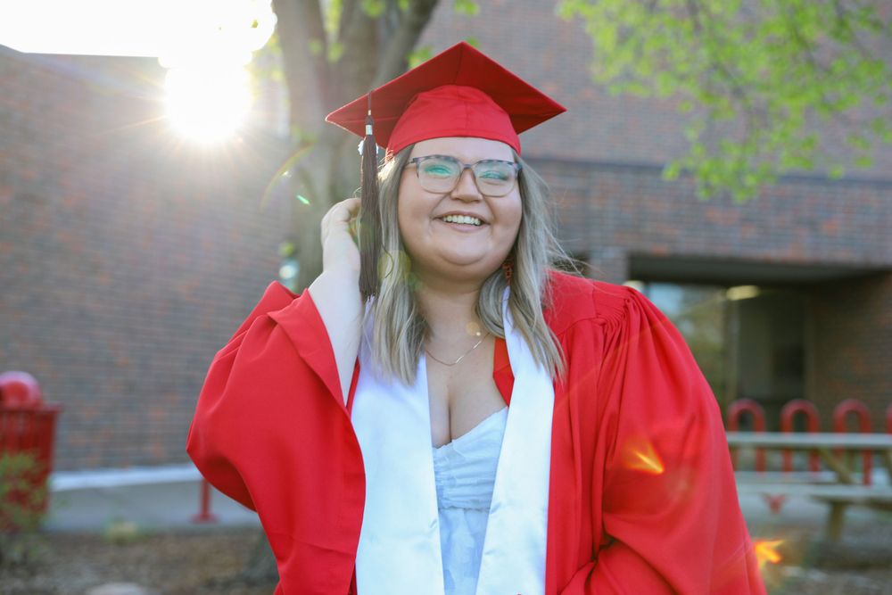 Graduate in bright red regalia smiles outdoors with sunlight and buildings in background.