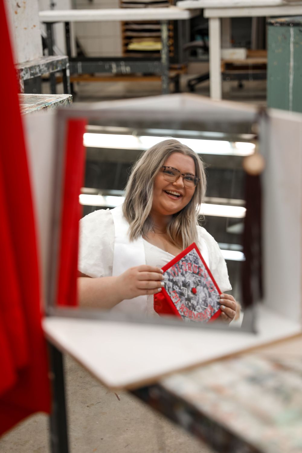 Graduate laughs while looking at decorated cap in classroom setting.