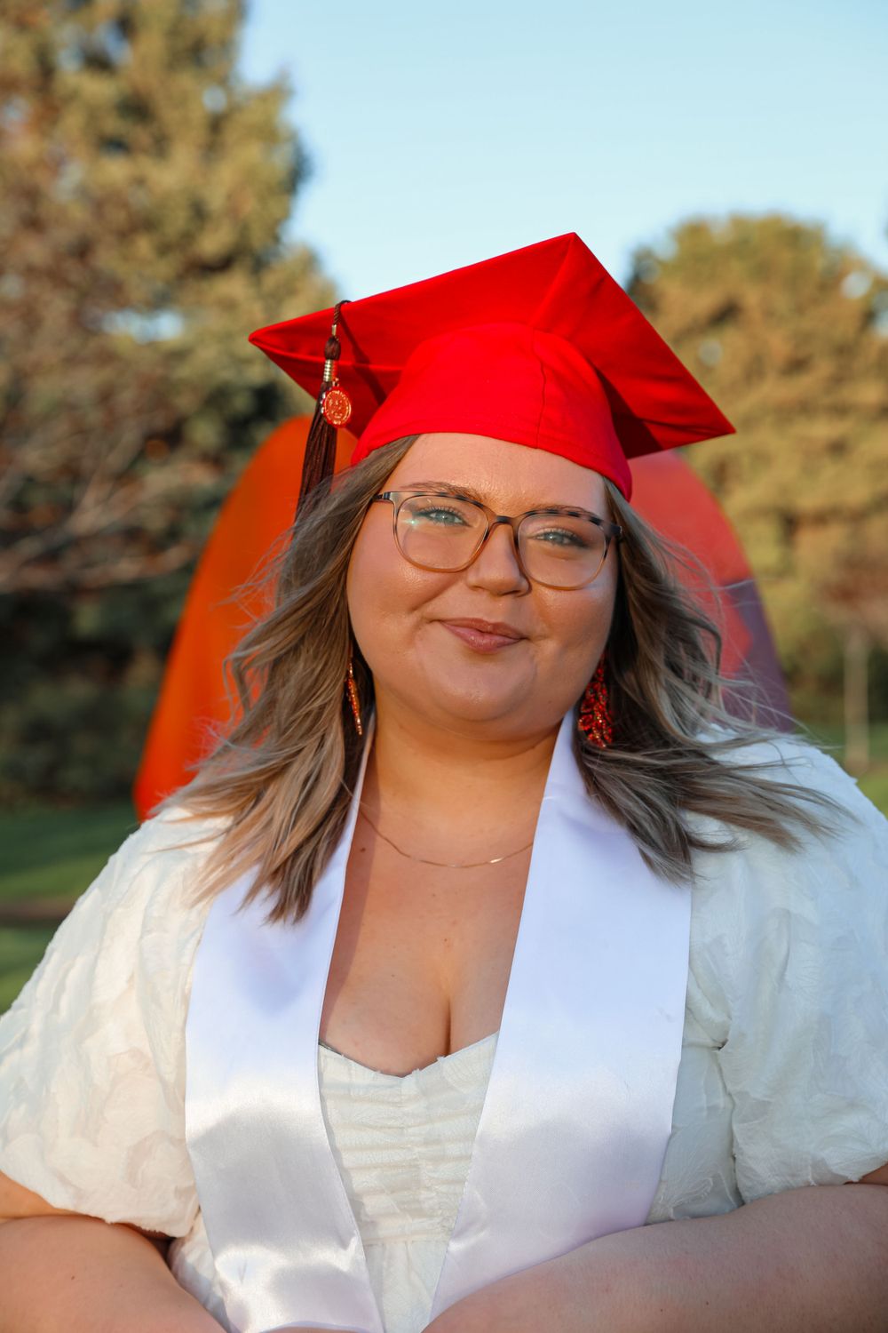 Graduate in traditional red cap and white sash poses during golden hour outdoors.
