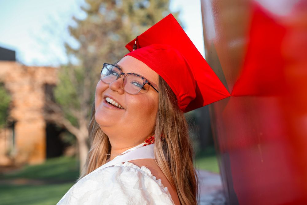 Graduate in red cap laughs joyfully against sunny outdoor backdrop.