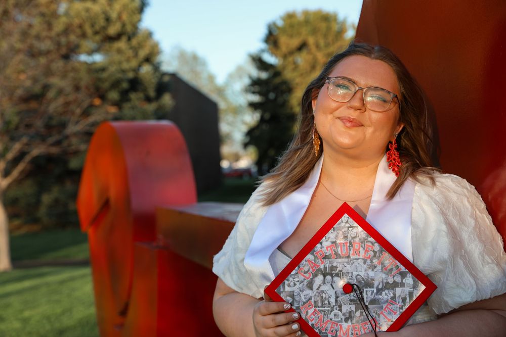 Graduate holds decorated cap while posing outdoors in traditional graduation attire.